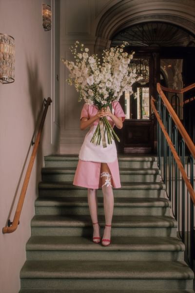 Woman in a pink maid dress holding a large bouquet of white flowers, standing on a staircase at Hotel Motto Vienna