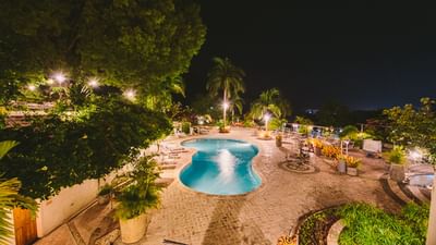 Illuminated pool by stone patio, surrounded by palm trees and string lights at night at Hotel Montana Haiti