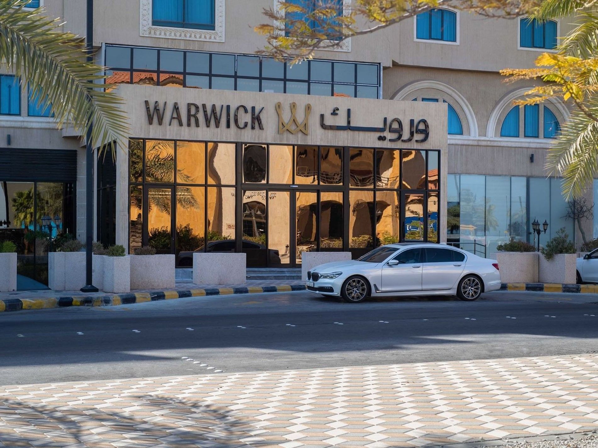 Modern entrance of Warwick Al Jubail Hotel with glass doors by stone planters under a large sign near a white sedan