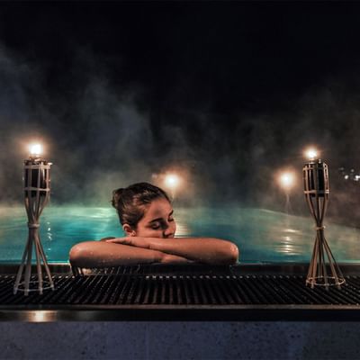 A woman relaxes in a hot tub at night, with her arms crossed and lit candles nearby.