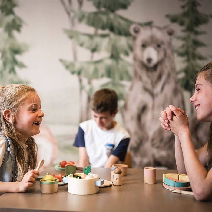 Kinder essen an einem Tisch mit Holzspielzeug und Wald-Bär-Wanddekoration bei Falkensteiner Hotel