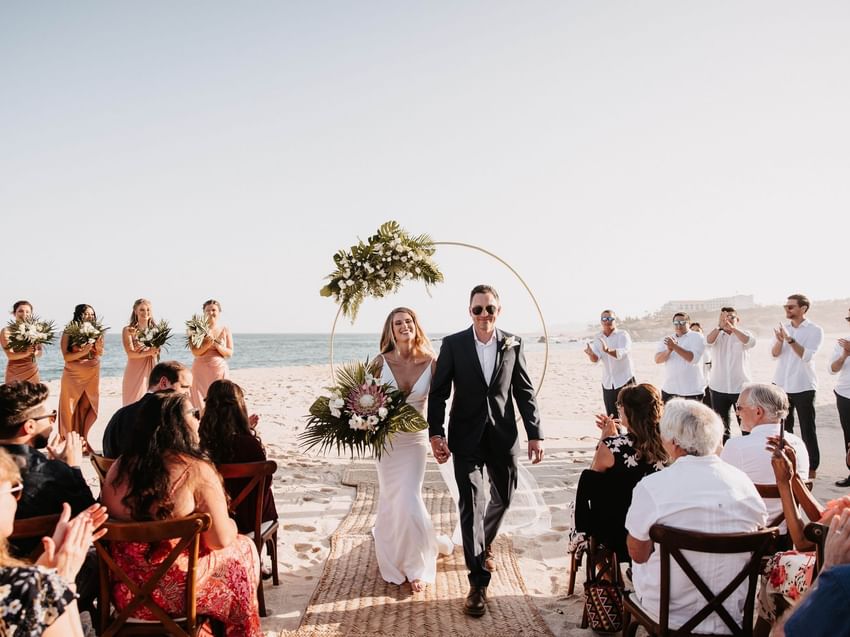 A newly married couple walks down a sandy aisle at their beach wedding at Marquis Los Cabos Resort guests applauding