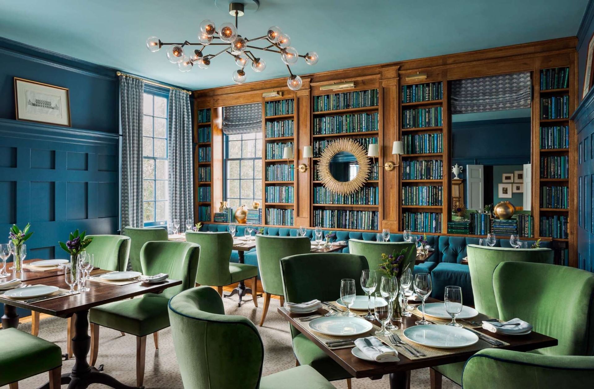 Green comfy chairs, dining tables with glassware, bookshelves & chandelier in The Library at The Clifton