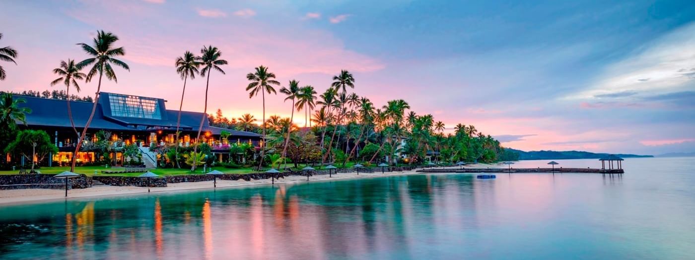 Tropical beach scene at sunset with palm trees, a serene ocean, and a beautiful The Warwick Fiji illuminated by warm lights