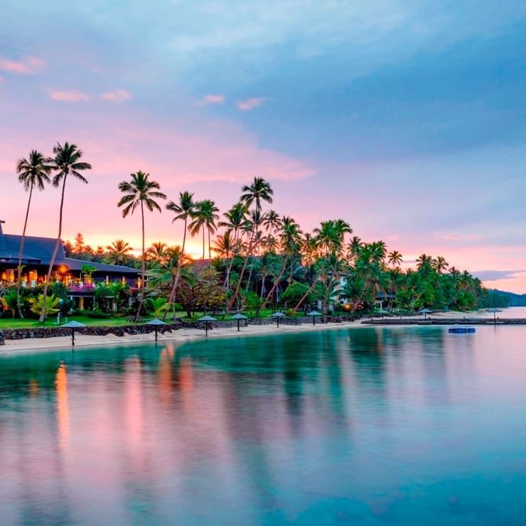 Tropical beach scene at sunset with palm trees, a serene ocean, and a beautiful The Warwick Fiji illuminated by warm lights