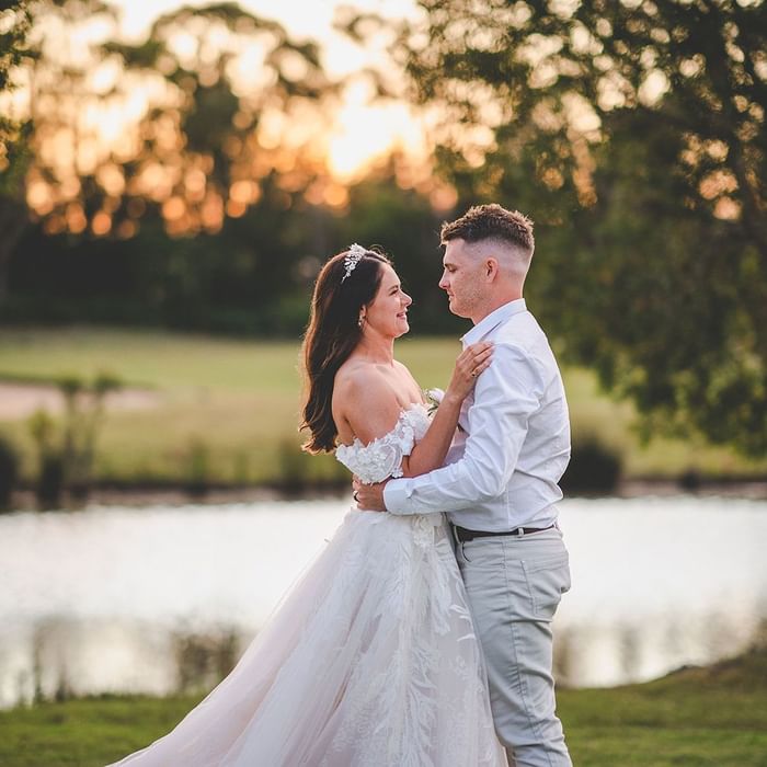 A bride and groom embrace by a lake at sunset, surrounded by trees near Mercure Kooindah Waters