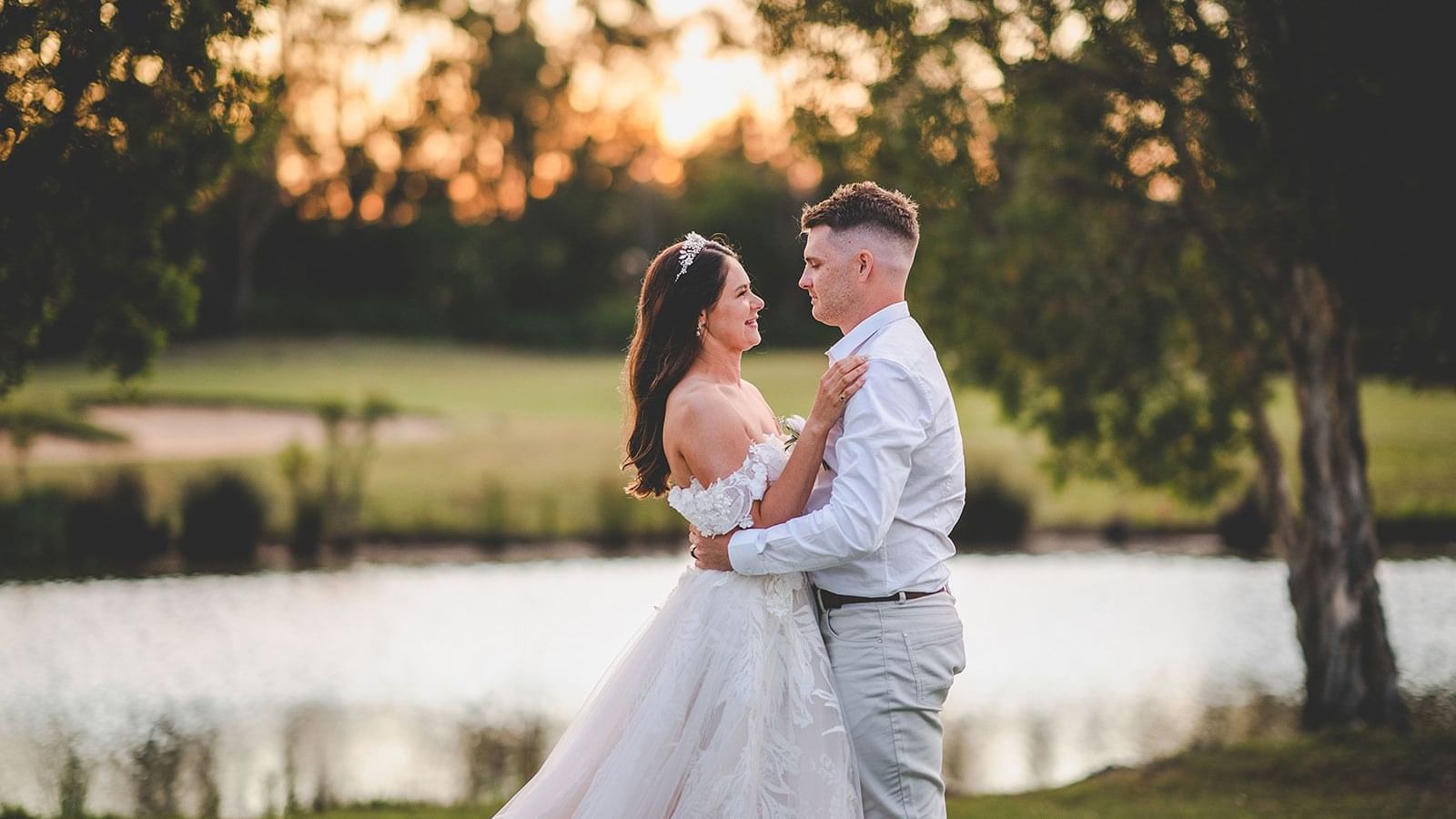 A bride and groom embrace by a lake at sunset, surrounded by trees near Mercure Kooindah Waters