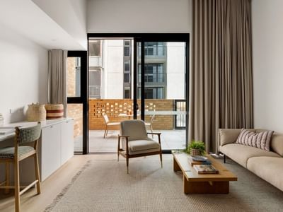 Modern living room with beige sofa, wooden coffee table, desk area, and balcony access through black-framed glass doors