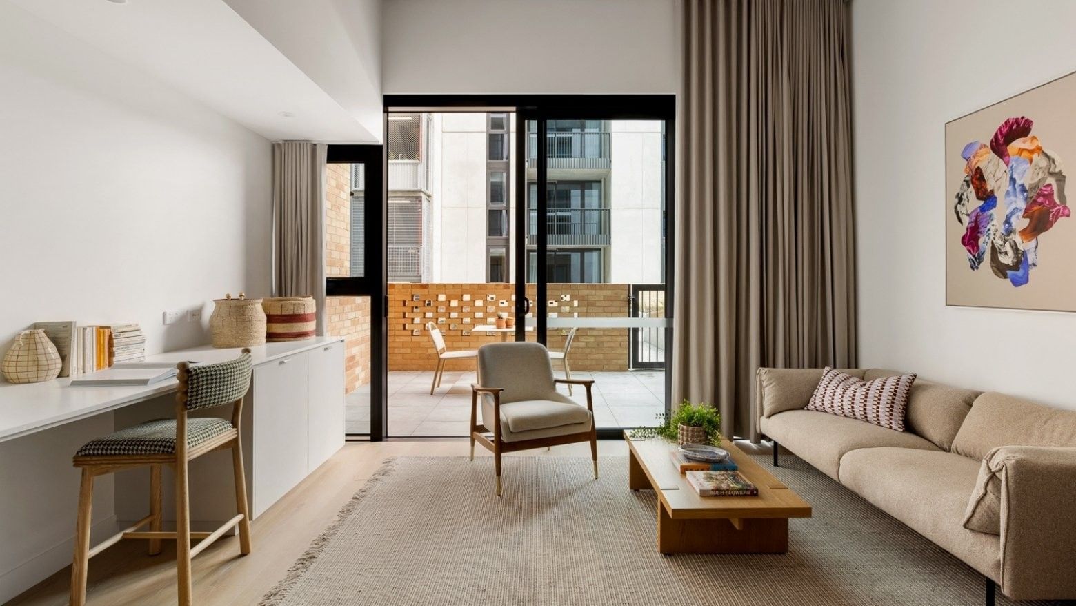 Modern living room with beige sofa, wooden coffee table, desk area, and balcony access through black-framed glass doors