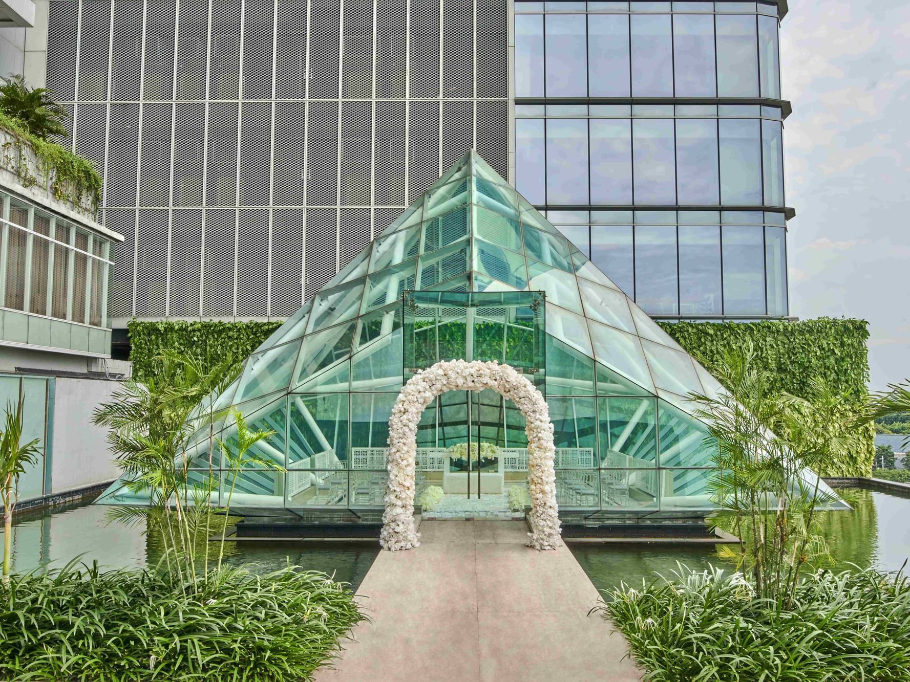 A glass pyramid structure with a floral arch, surrounded by greenery and water at Vasa Hotel Surabaya