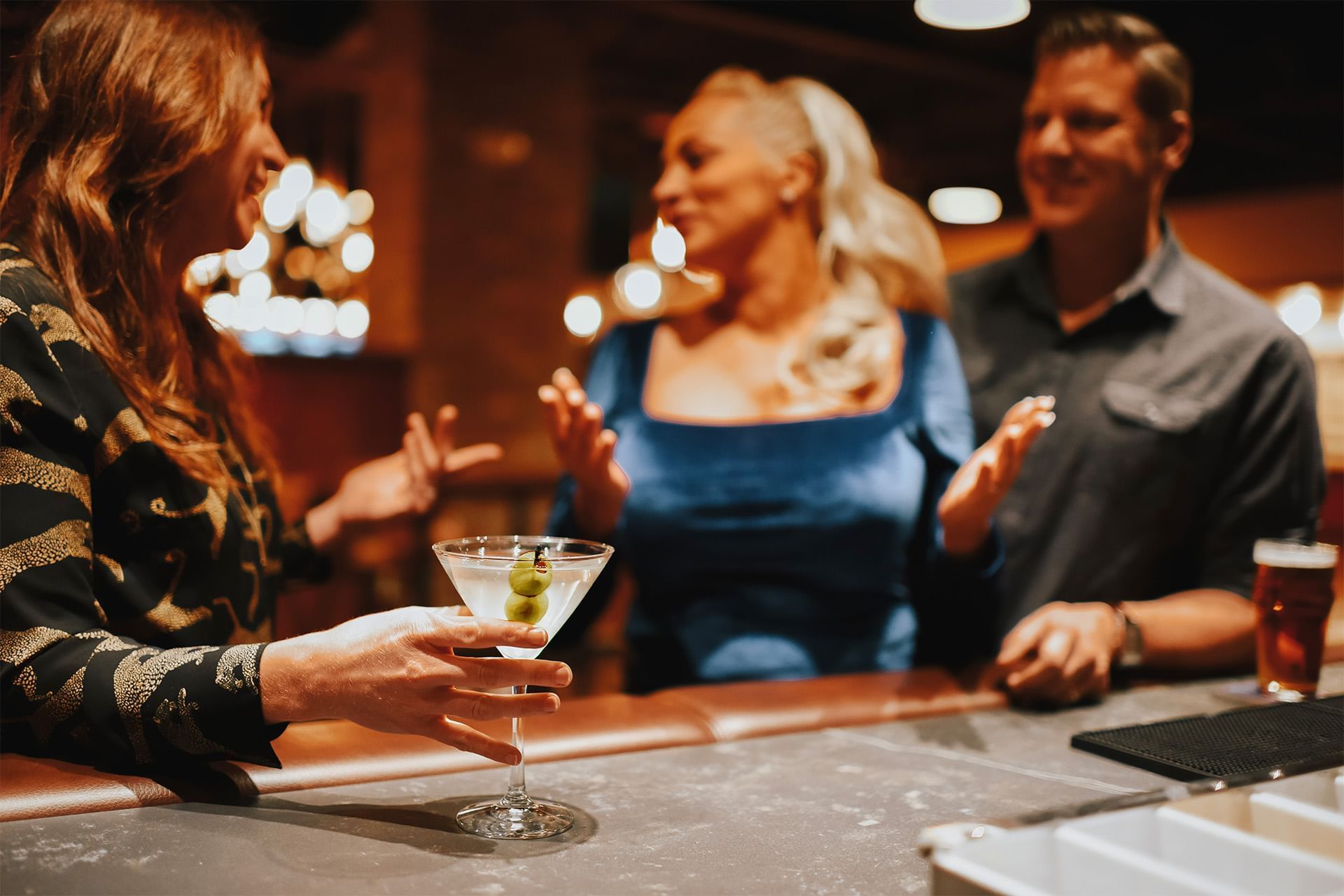 Friends chatting by a martini glass with olives and a beer on a marble bar counter under warm chandeliers at The Brook