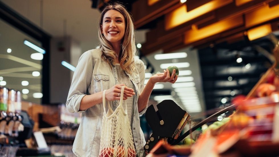 Woman holding a green apple and a reusable bag filled with produce in a grocery store.