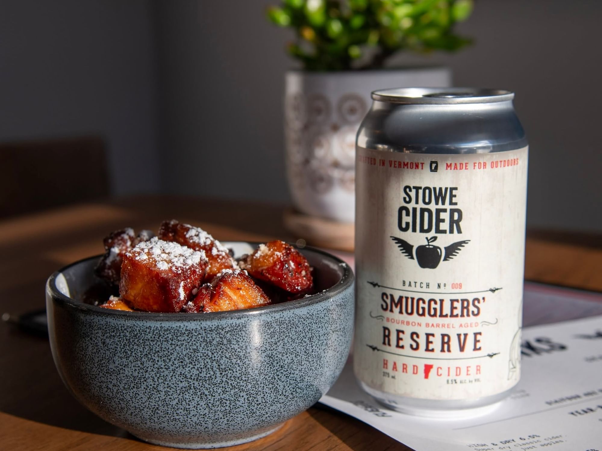 Can of Stowe Cider Smugglers' Reserve hard cider beside a bowl of powdered sugar-dusted pastries on a wooden table.