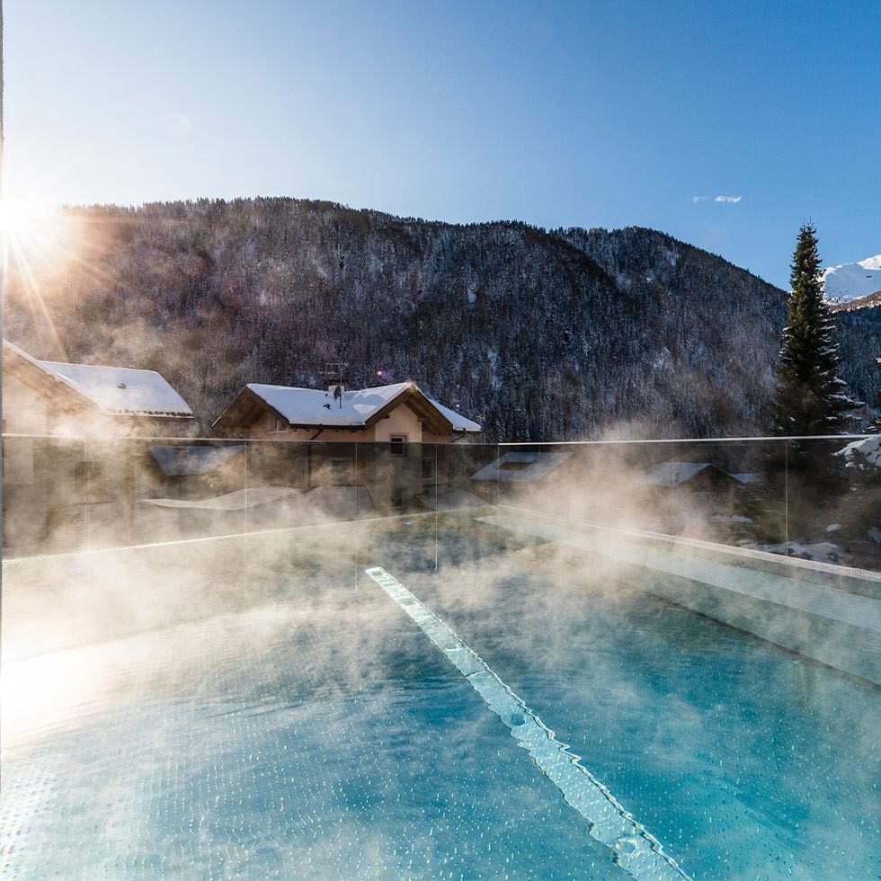 Piscina esterna riscaldata con vapore, montagne innevate sullo sfondo, sole splendente.