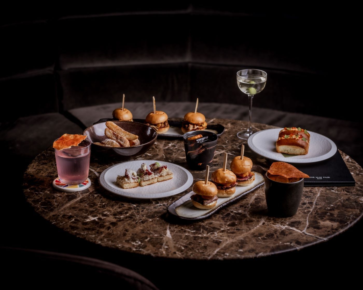Assorted appetisers and cocktails from the by night menu on a round marble table at The May Fair Bar