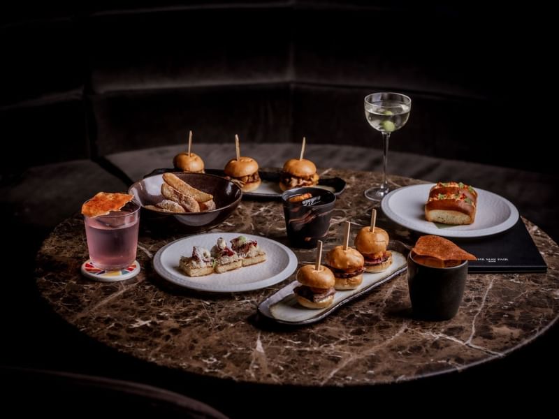 Assorted appetisers and cocktails from the by night menu on a round marble table at The May Fair Bar