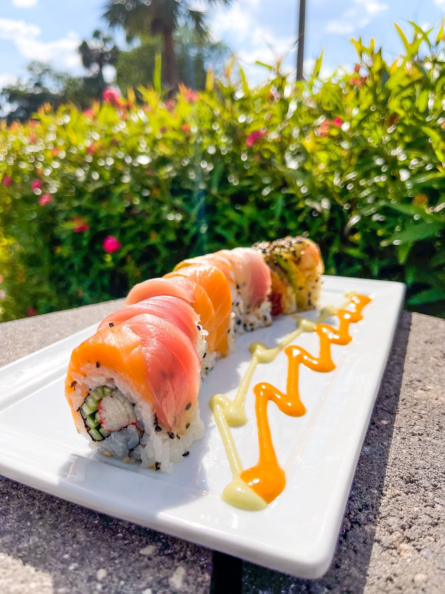 A curving sushi roll topped with colorful fish on an outdoor table in front of pink flowering shrubs,