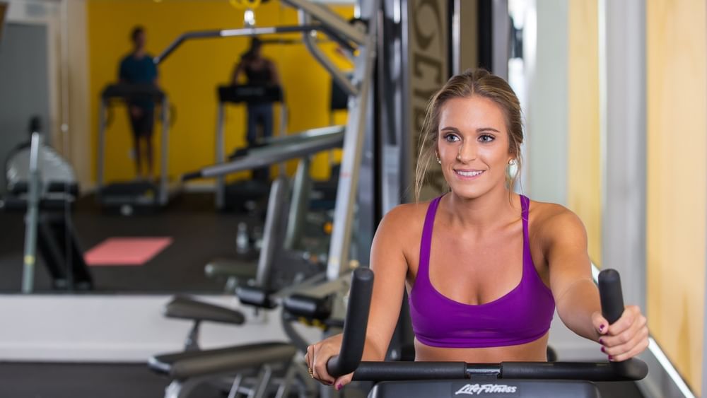 Woman on a bike by gym mirrors under bright light surrounding fitness equipment at Warwick Paradise Island Bahamas