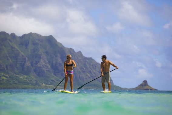 Couple paddleboarding with a mountain backdrop on a sunny day near Paradise Bay Resort