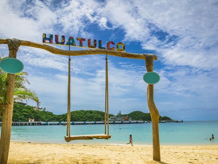 Columpio de madera en la playa bajo letrero “Huatulco” con agua turquesa tranquila cerca de Camino Real Huatulco