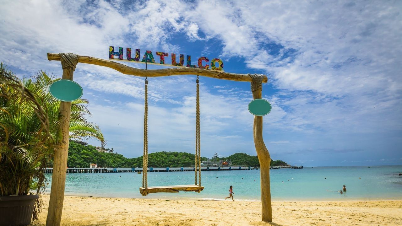 Columpio de madera en la playa bajo letrero “Huatulco” con agua turquesa tranquila cerca de Camino Real Huatulco