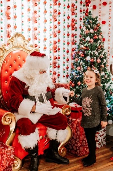 A girl in a sweater greets Santa in front of a wall of ornaments and a decorated Christmas tree at Wekiva Island's Winter Wonderland, a great place to meet Santa in Orlando