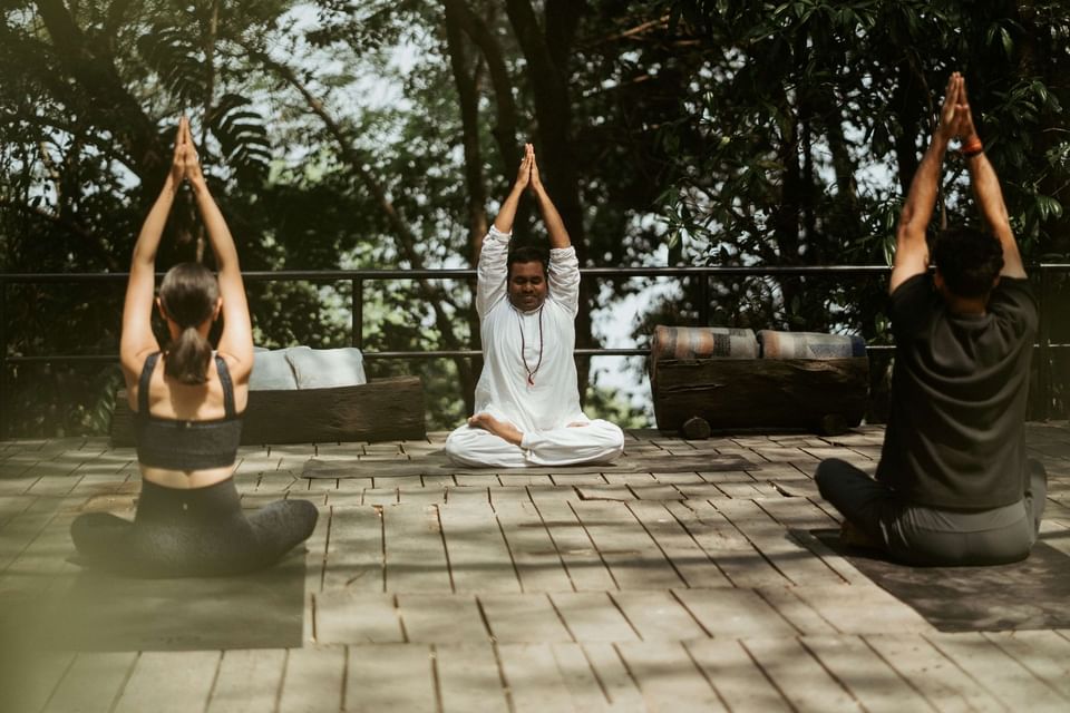 Three people practice yoga on a wooden deck surrounded by greenery at The Dwarika's Himalayan Wellbeing