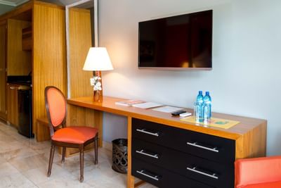 Wall-mounted TV above a wooden desk with water bottles by an orange chair in Superior Room at Hotel Montana Haiti