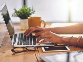 A person typing on a laptop with a coffee cup and small potted plant on a desk.