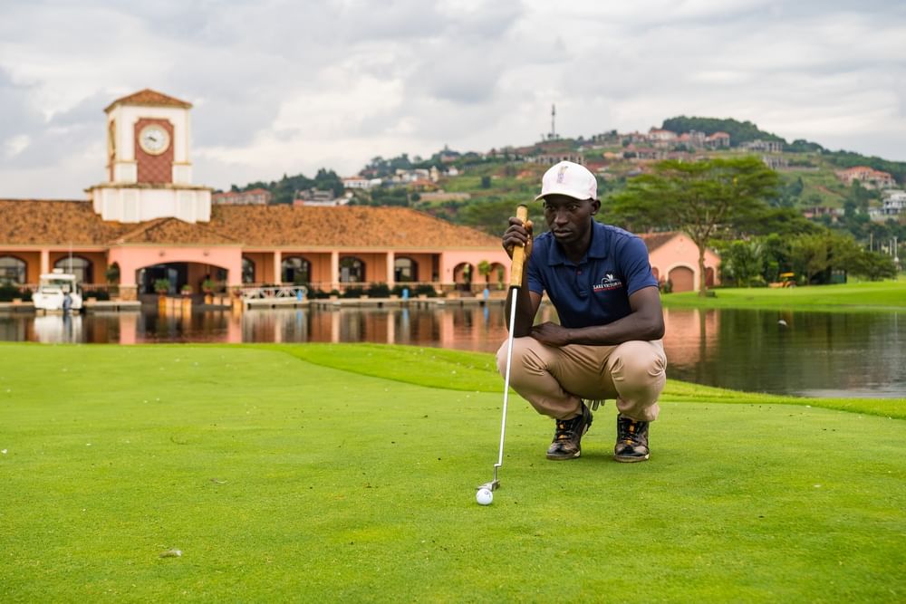 Golfer crouching on green with flag and ball beside water feature and terracotta buildings at Kampala Serena Hotel