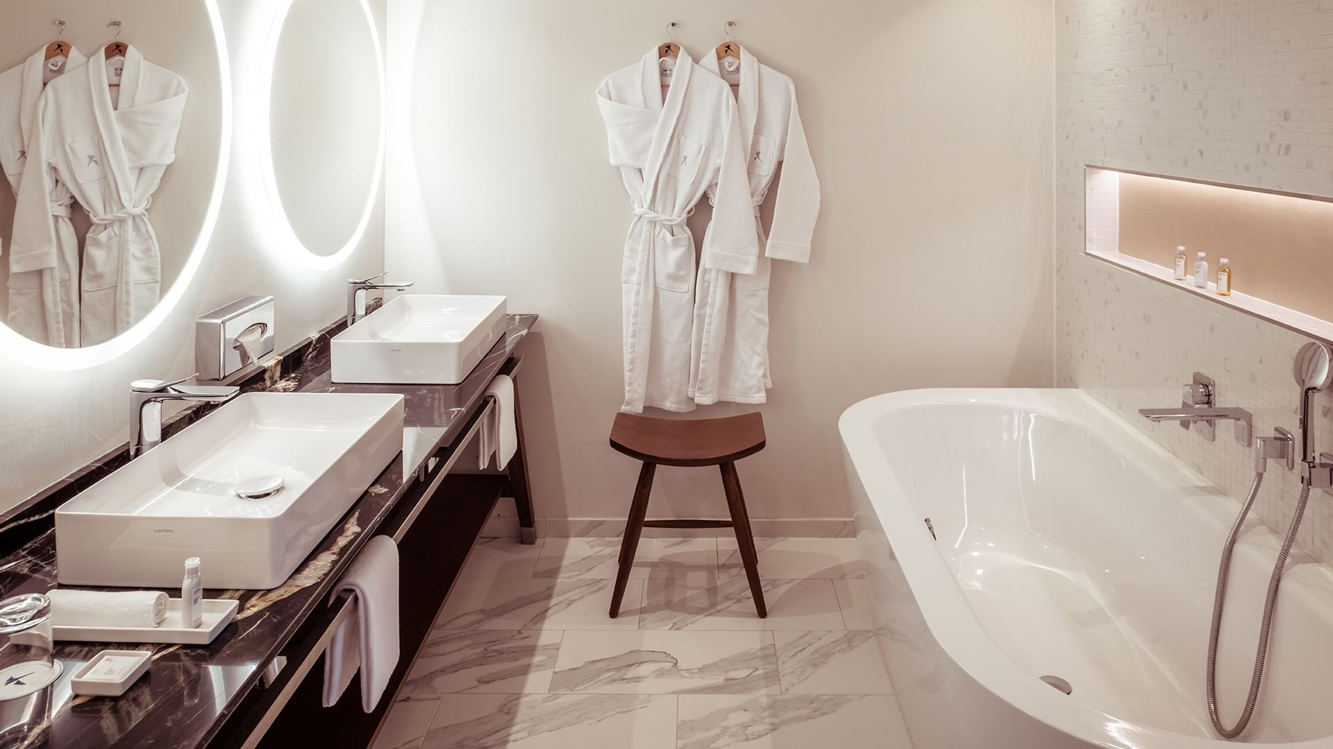 Elegantes Badezimmer mit zwei Waschbecken, beleuchteten Spiegeln, freistehender Badewanne und hängenden Bademänteln.