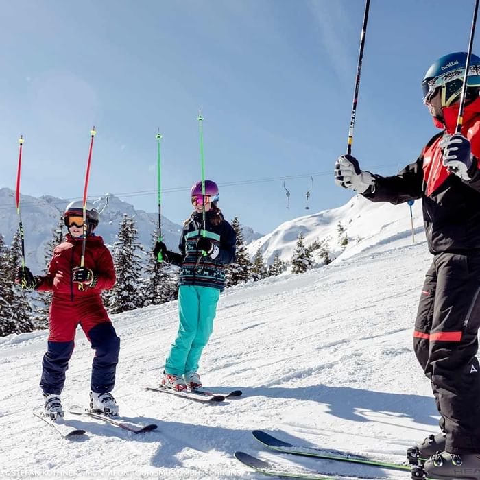 Tre sciatori con istruttore su una pista da sci in una giornata di sole, montagne innevate sullo sfondo.