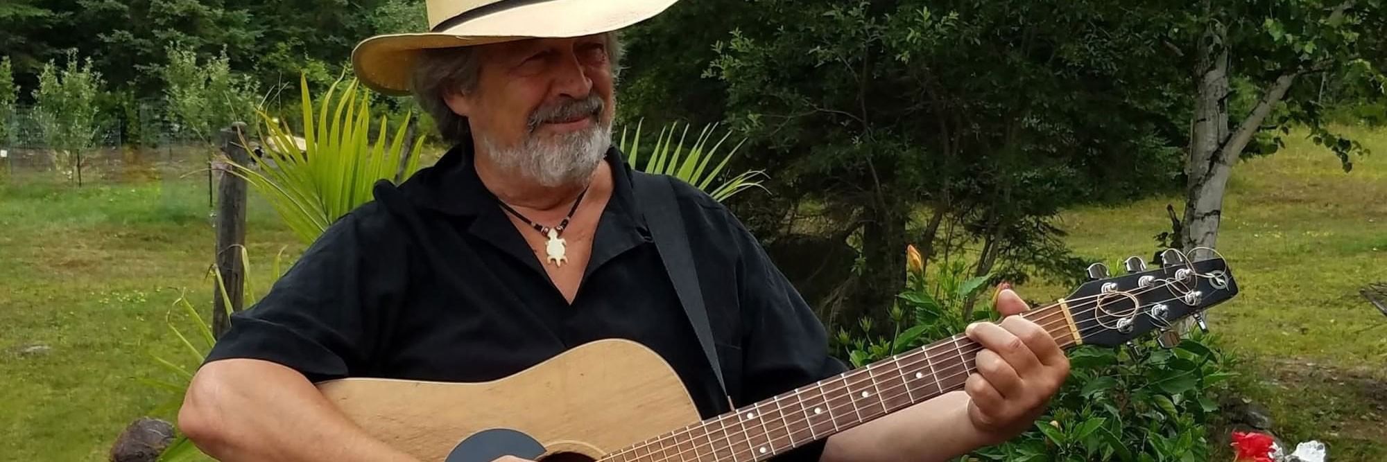 Wayne Failing in a cowboy hat playing a guitar by a wooden fence, surrounded by green garden plants.