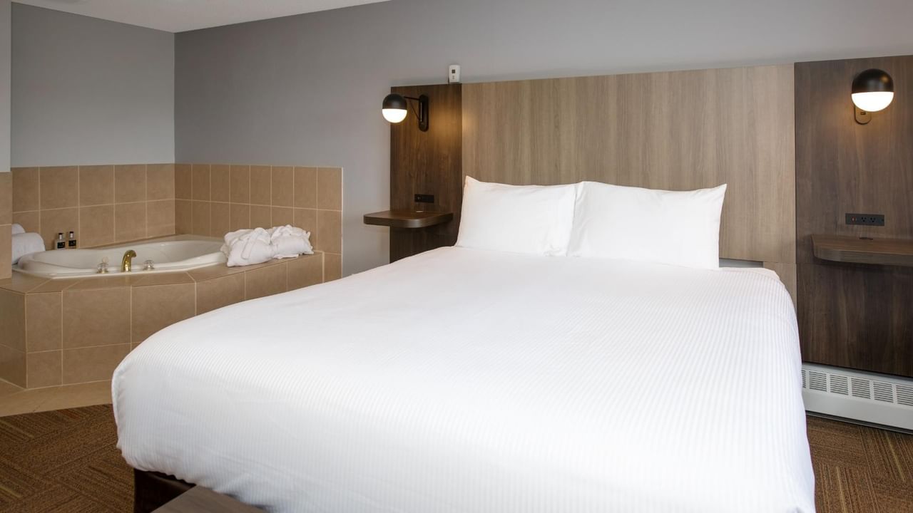 A hotel room with a large bed, two white pillows, and a bathtub with towels.