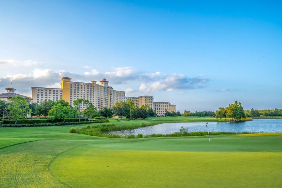 A large expanse of green grass on a golf course with a golf flag overlooking a lake, trees, and a towering hotel in the distance.