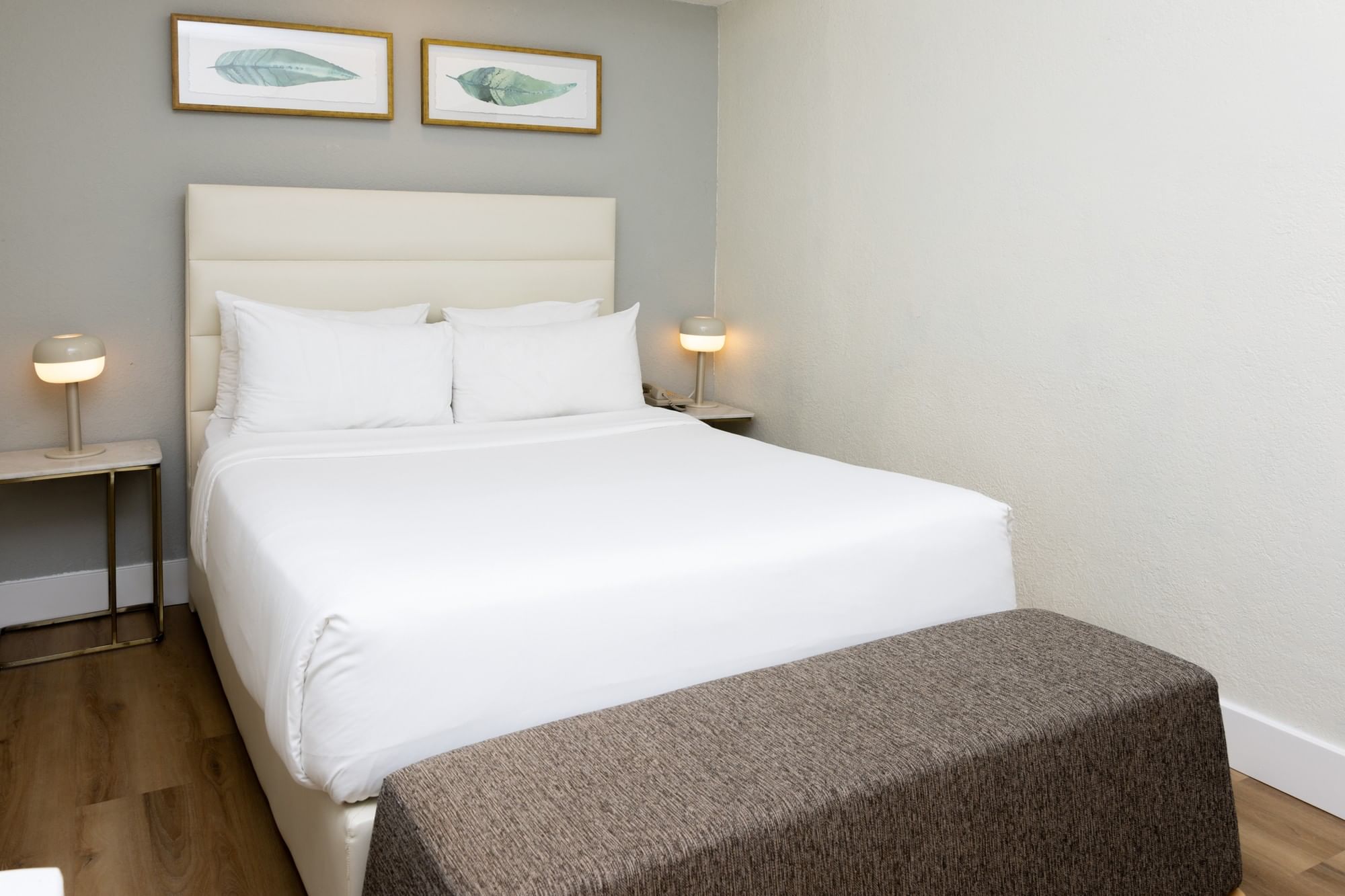 A neatly made bed with white linens and pillows, flanked by two nightstands with lamps, and a bench at the foot of the bed, in a room with gray walls and wood floors.