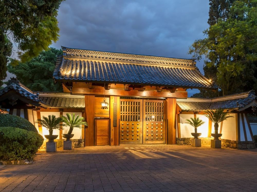Lit traditional Japanese-style gate with a tiled roof and wooden doors, surrounded by trees at Hotel Sumiya Cuernavaca