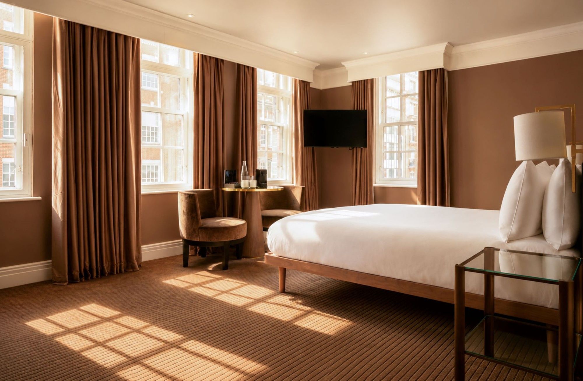 Sunlit Collection Deluxe King Room with brown curtains, white bedding, desk, and wall-mounted TV at The May Fair Hotel, London