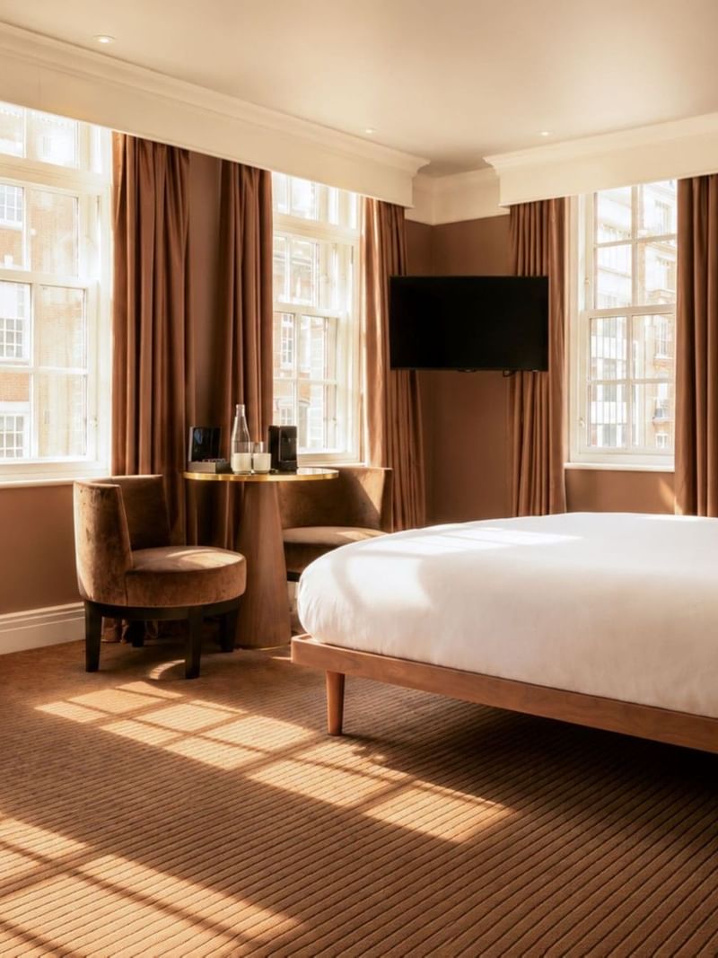 Sunlit Collection Deluxe King Room with brown curtains, white bedding, desk, and wall-mounted TV at The May Fair Hotel, London