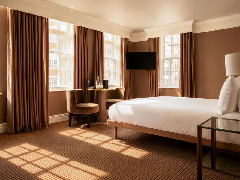Sunlit Collection Deluxe King Room with brown curtains, white bedding, desk, and wall-mounted TV at The May Fair Hotel, London