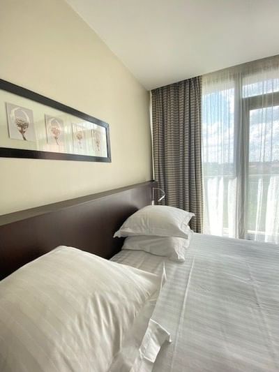 Lit avec trois oreillers et une tête de lit en bois sous une image encadrée près de fenêtres.