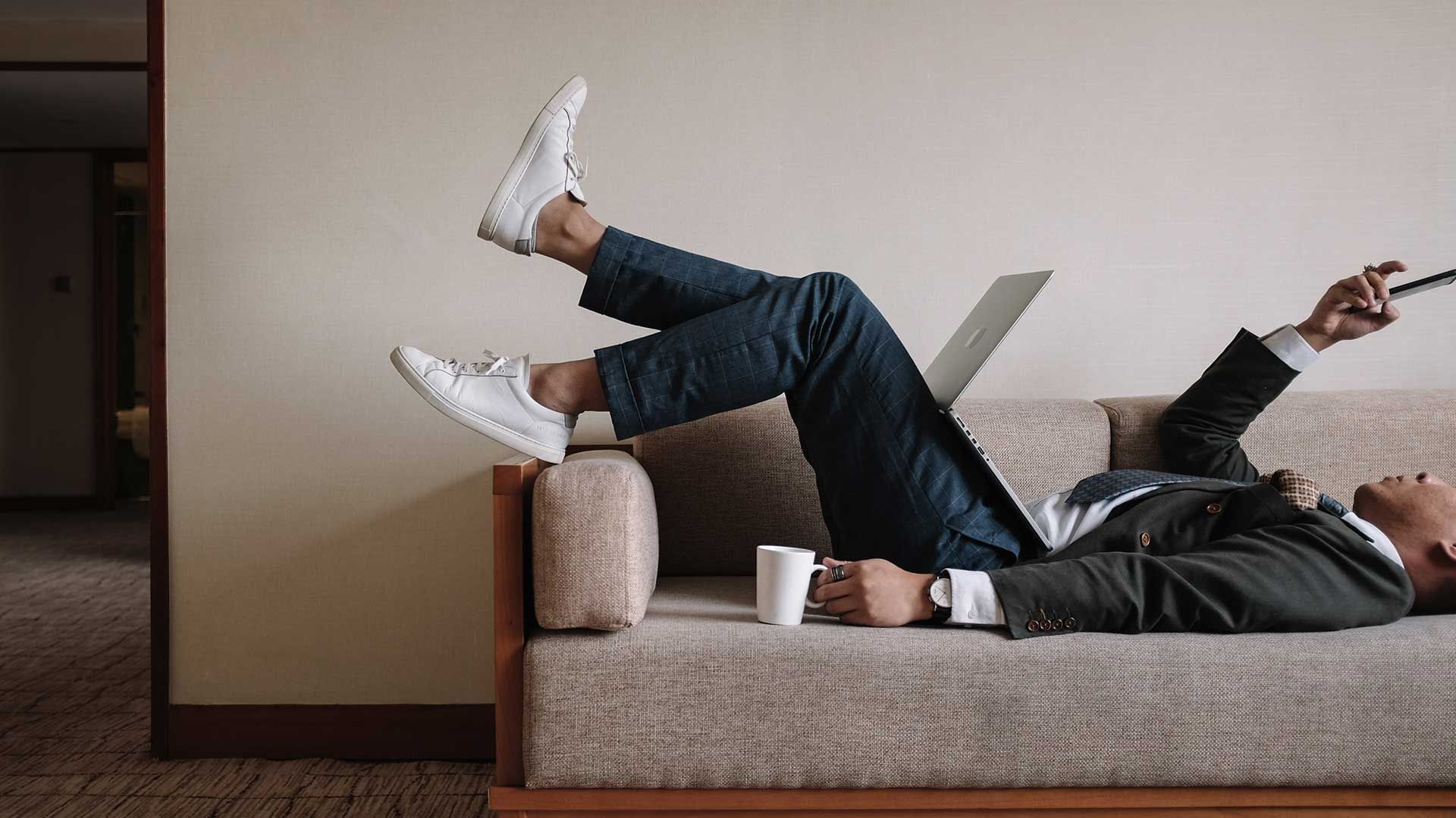 A man laying on a sofa with coffee, laptop and phone at Park Hotel Group
