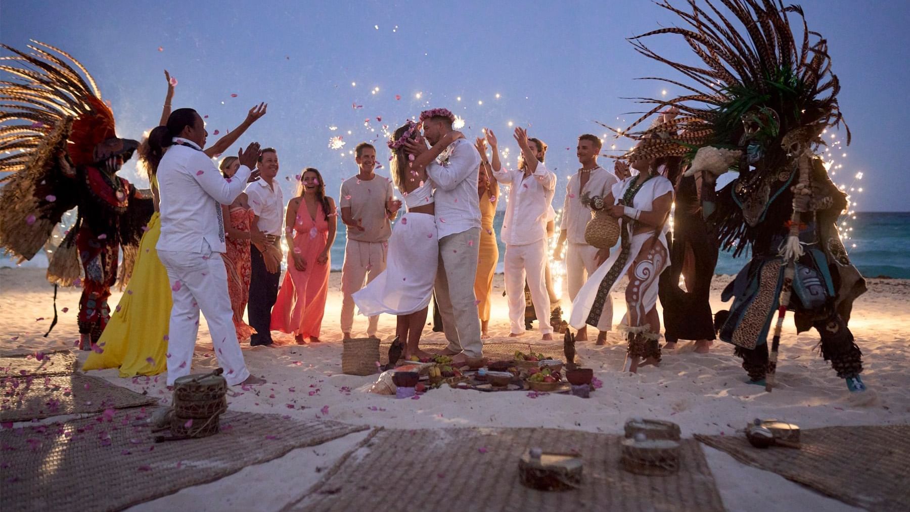 A group of people celebrating with a couple on a beach at dusk near Grand Fiesta Americana