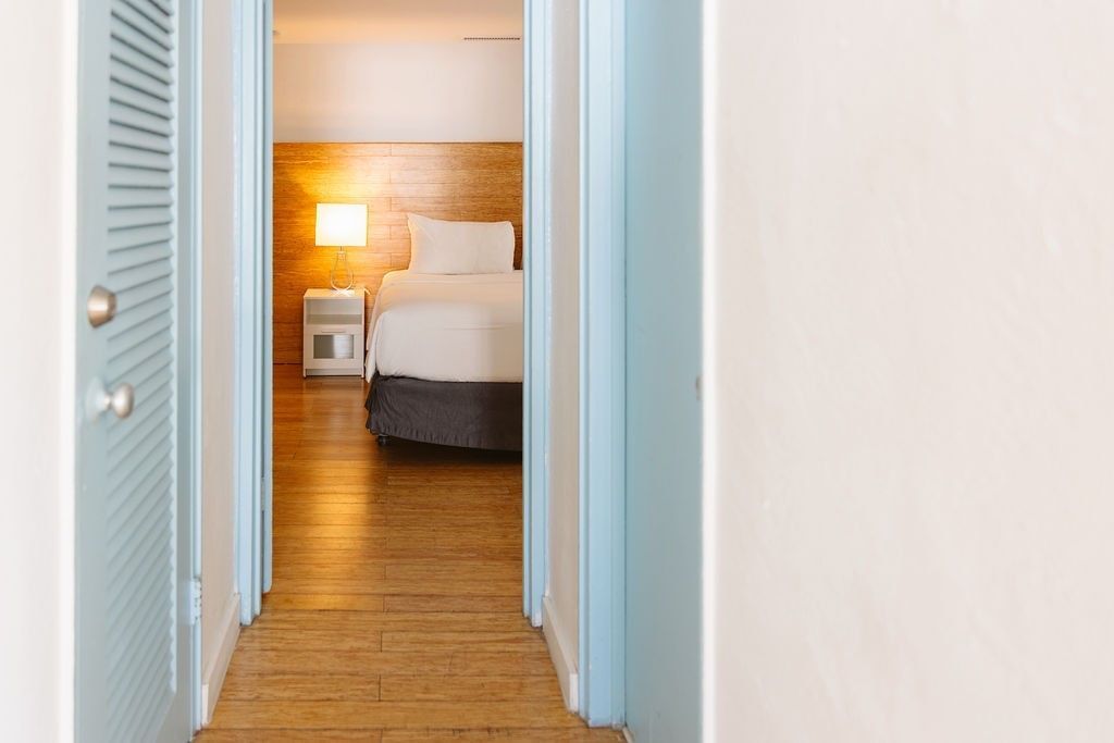A hotel room viewed through an open doorway, with a bed, nightstand, and lamp visible.