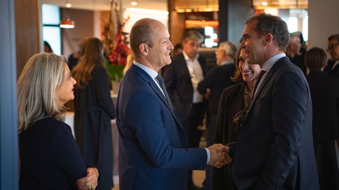 Two men shake hands in conversation at a social event, surrounded by guests and floral decor at Oceania Hotels