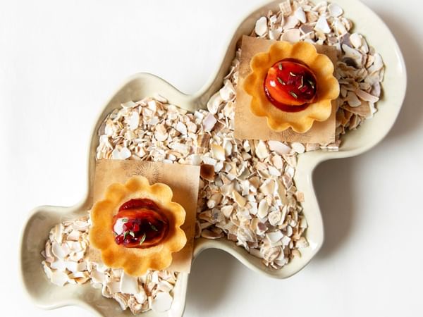 Two tarts with fruit fillings on a heart-shaped plate filled with shredded coconut on a white background.