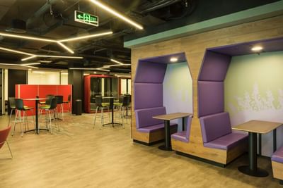 Modern office space with purple booths, wooden accents, and a casual dining area with tall bar tables & stools at Hotel Nodo