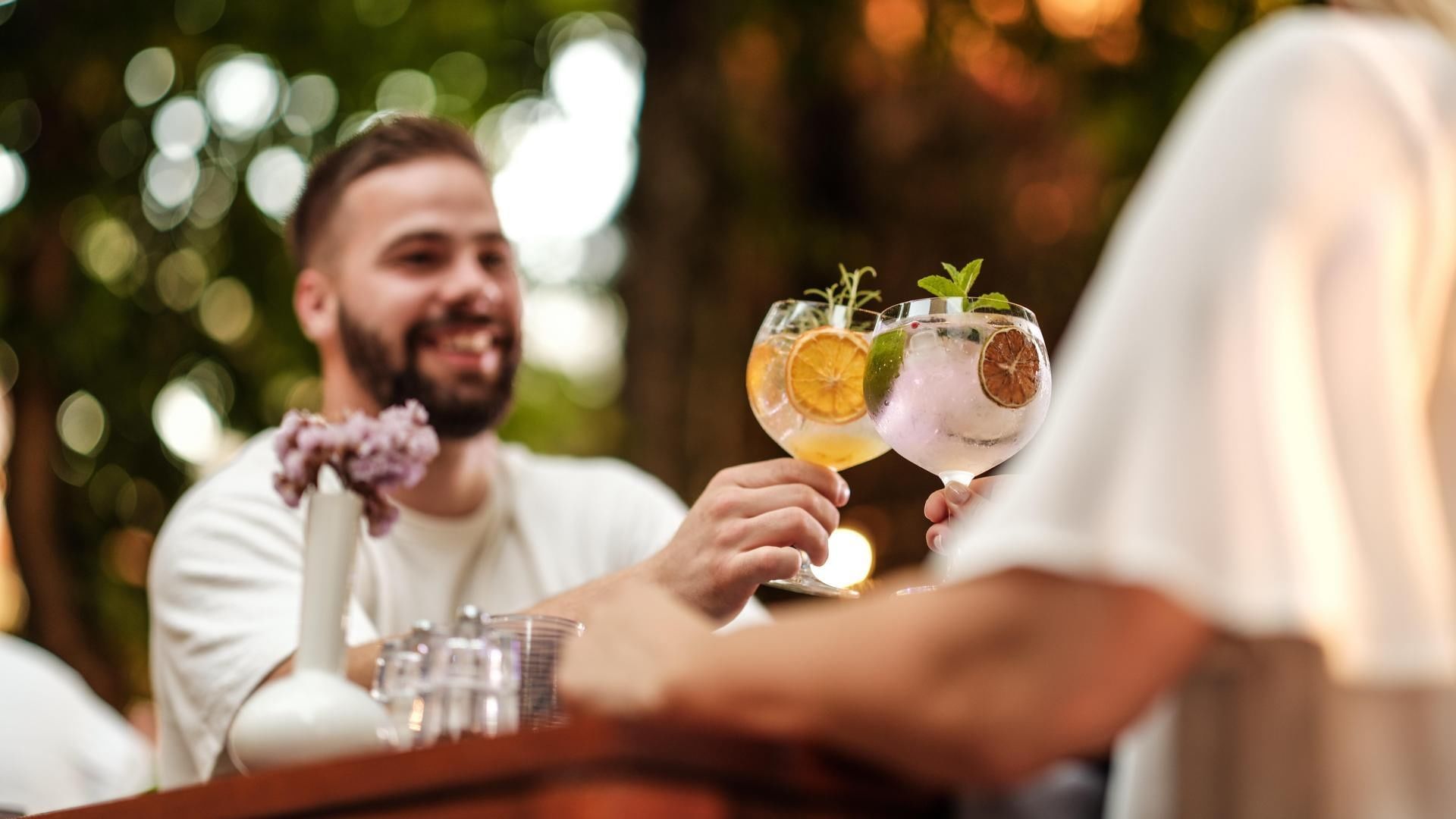 Two people holding cocktails with garnishes and a vase with flowers on a table.