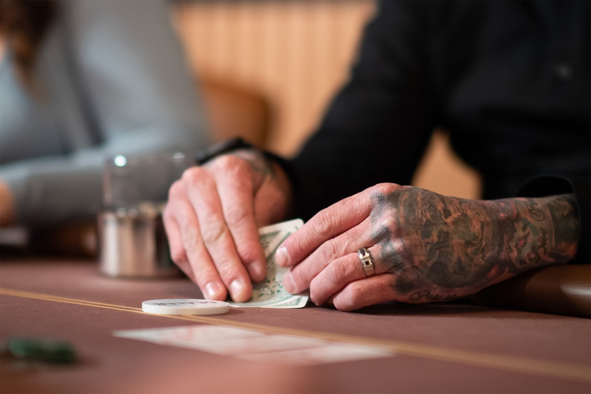 Tattooed dealer\'s hand sliding a card across smooth burgundy felt, beside a poker chip and metal shaker at The Brook