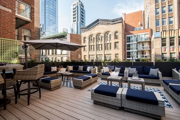 Blue sofas surrounding wicker tables by a grey umbrella on the wooden rooftop deck at HGU New York Hotel